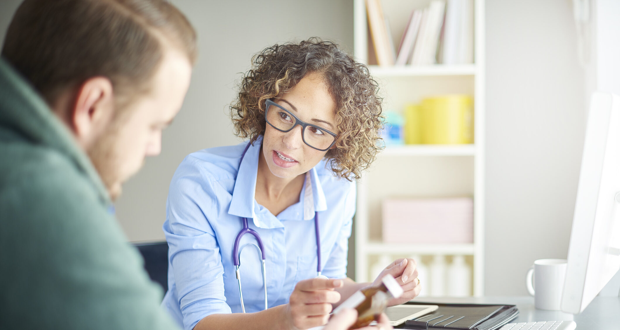 A female doctor sits at her desk and chats to a male patient about his current medication . She is dressed in a shirt with rolled up sleeves . They are both looking down at the pill bottle as she assesses his current dosage.